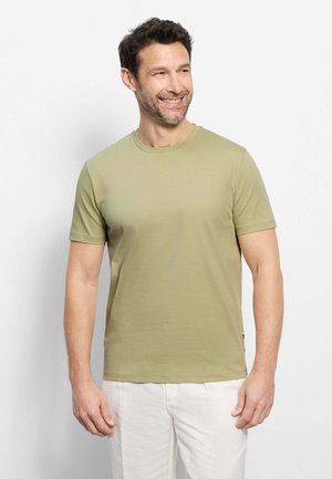 Lachende man met kort donker haar en baard, gekleed in een lichtgroen T-shirt met korte mouwen en witte broek, kijkend naar rechts.