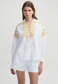 Camicia bianca a maniche lunghe con motivi ricamati gialli, colletto a V, cordoncino e bottoni frontali. Indossata con pantaloni corti bianchi.