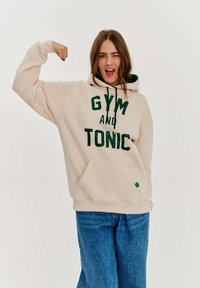 Beige mikina s kapucí a zeleným nápisem "GYM AND TONIC". Má klokaní kapsu, kapuci se šňůrkou a je spárována s modrými džíny.