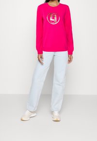 Fuchsiová mikina s dlouhými rukávy a kulatým výstřihem, se velkým zlatým logem ve středu, spárovaná s světle modrými džíny a béžovými teniskami.