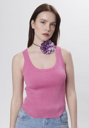 Roze geribbelde tanktop met een diepe halslijn, gecombineerd met een grote paarse bloemenaccessoire op een zwarte choker. Eenvoudige lichte achtergrond.