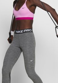 Różowy stanik sportowy z magentową taśmą i czarnym logo Nike; szare dopasowane legginsy z czarnym elastycznym pasem z białym napisem "NIKE PRO".