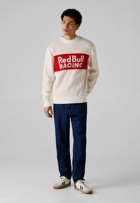 Kreminės spalvos megztinis su raudonu horizontaliu dryžu, ant kurio baltomis raidėmis užrašyta „Red Bull RACING“, derinamas su tamsiai mėlynais džinsais ir baltais sportbačiais.