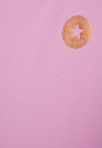 Vaaleanpunainen puuvillainen t-paita, jossa on pyöreä, kohokuvioitu, tan-nahkainen merkki, jossa lukee "Converse All Star" ja tähtilogo. Sileä pinta, pyöreä kaula-aukko.