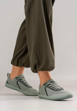 Hellgrüne Netzsneaker mit strukturierten Details, einer flexiblen Sohle und Schnürverschluss, kombiniert mit lockeren, dunkelgrünen, taillierten Hosen.