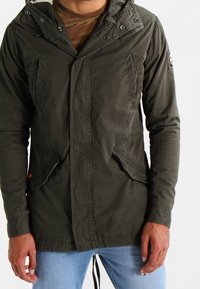 Olive green jacket featuring a hood, front snap closures, two side pockets, and a drawstring hem, made from a smooth, durable fabric.