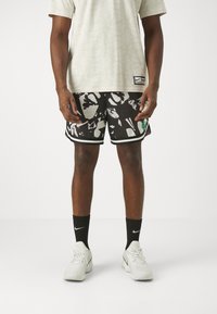 Pantalones cortos de baloncesto de malla en blanco y negro con un patrón floral, que cuentan con un logo verde y ribete negro. Se llevan con una camisa de patrón de color claro y zapatillas blancas.