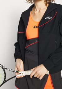 Svart jacka med röda detaljer, med dragkedja framtill och elastiska ärmar, bärs över en orange topp. Person som håller en tennisracket.