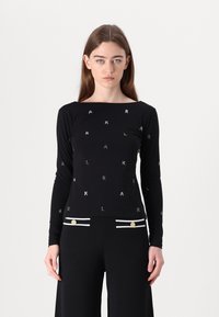 Schwarzes Langarmtop mit quadratischem Ausschnitt und verstreuten silbernen Buchstaben. Getragen mit schwarzen Hosen, die mit weißen Streifen und goldenen Akzenten verziert sind.