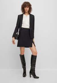 Schwarzer Blazer über einer weißen Bluse, kombiniert mit einem schwarzen Minirock mit einem seitlichen Schlitz. Knielange, glänzende schwarze Stiefel mit Absatz runden das Outfit ab.