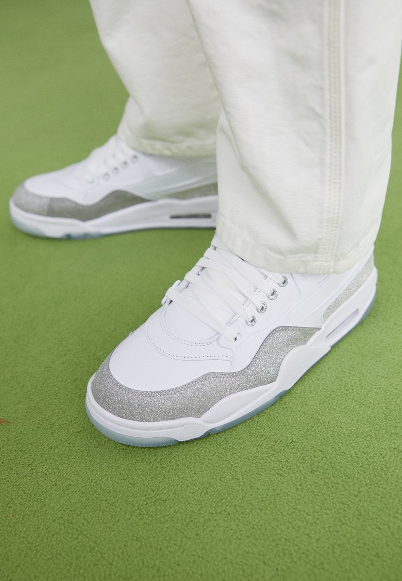 Sneakers bianchi con accenti di glitter argentati, design con lacci, collare imbottito e suola traslucida, posizionati su una superficie verde.