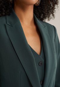 Mörkgrön kavaj med spetsiga slag och V-halsstil under. Har två svarta knappar och ett slätt, texturerat tyg.