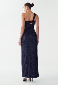 Viena pleca, tumši zils mirdzošs vakarkleita ar savītu siksnu, atvērumu aizmugurē un piegulošu siluetu. Redzamas sudraba krāsas augstpapēžu kurpes.