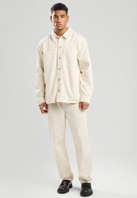 Light beige button-up shirt with two chest pockets, paired with matching trousers. Smooth fabric texture; worn with black shoes.