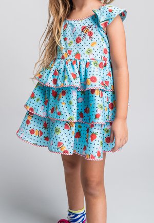 Młoda dziewczyna ubrana w jasnoniebieską sukienkę warstwową z kolorowymi wzorami owoców i bufkami, stojąca na jednolitym szarym tle.