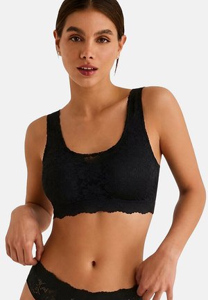 Femme portant un bralette en dentelle noire et une lingerie en dentelle assortie sur un fond clair uni.