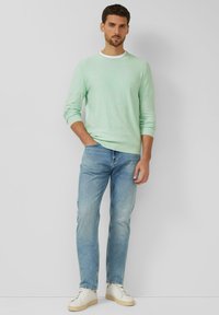 Man staat met hand in zak, draagt een mintgroene trui, blauwe spijkerbroek en witte sneakers tegen een effen lichte achtergrond.