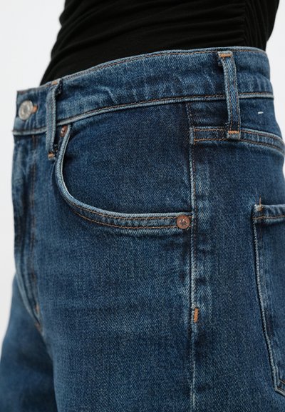 Modre kavbojke iz jeansa z značilnim klasičnim dizajnom s petimi žepi, bakrenimi okovji, subtilnimi šivi in rahlo teksturirano površino.