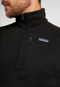 Schwarzer Fleece-Pullover mit Stehkragen, halber Reißverschluss und einer Brusttasche. Verfügt über ein kleines blaues und weißes Patagonia-Logo. Weiche Textur.