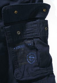 Granatowy materiał z załataną kieszenią, wyposażoną w żółte i srebrne metalowe zatrzaski. Nadrukowana, vintage etykieta w kolorach niebieskim i beżowym z dekoracyjnymi gwiazdkami.