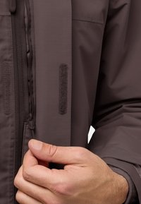 Bruine waterdichte jas met een ritssluiting aan de voorkant en klittenbandaccenten. Heeft een gestructureerde, lichte stof en is verstelbaar met een trekkoorden.