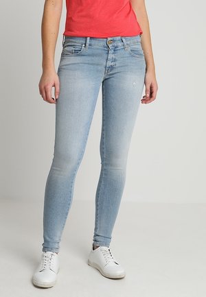 Jeans ajustados de color azul claro con un acabado desgastado, diseño de cinco bolsillos y cierre de botón metálico, combinados con zapatillas blancas.