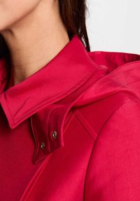 Giacca rossa con colletto prominente e dettagli sui bottoni, in tessuto liscio, e cappuccio integrato nel design, che mette in risalto una silhouette elegante.