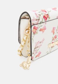 Biała torebka w kwiatowy wzór z złotym łańcuszkiem i zawieszką w kształcie królika, charakteryzująca się usztywnioną konstrukcją i teksturowanym wykończeniem.