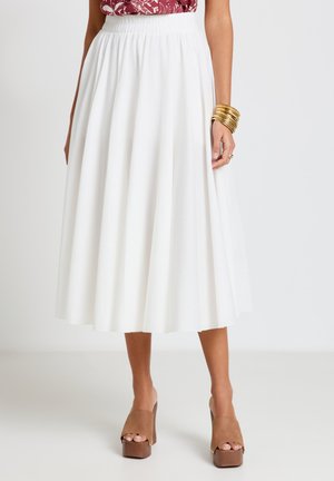 Femme portant une jupe midi blanche avec une taille élastique, associée à des sandales à plateforme marron à bouts ouverts et à des bracelets dorés au poignet.