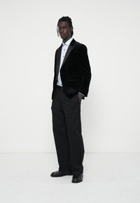 Blazer in velluto nero con rever in satin, abbinato a una camicia azzurra chiara e pantaloni neri. Il modello sta in piedi con sicurezza con le mani nelle tasche.
