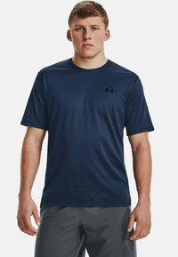 Jaunas vyras su trumpais plaukais, vilkintis tamsiai mėlyną Under Armour marškinėlį ir pilkas šortus, stovi prie paprasto šviesaus fono.