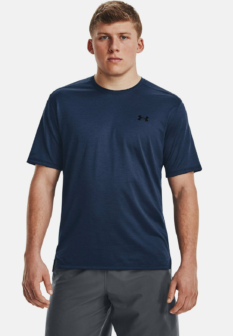 Jaunas vyras su trumpais plaukais, vilkintis tamsiai mėlyną Under Armour marškinėlį ir pilkas šortus, stovi prie paprasto šviesaus fono.