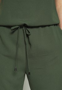 Gros plan sur un pantalon en tissu vert et un haut avec un cordon de serrage noué à la taille, doté d'embouts métalliques aux extrémités du cordon.