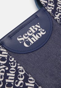 Zbliżenie na granatową tkaninę z białym logo "See By Chloé" na skórzanym metce oraz tekstylnym wykończeniu.