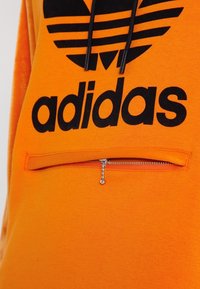 Orange Adidas hoodie med svart trefoil-logga, svarta dragskor och en horisontell dragkedjeficka med silverhandtag.