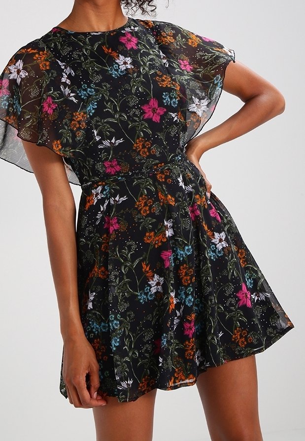 Kukallinen mekko mustalla läpikuultavalla kerroksella, jossa on värikkäitä kukkia vaaleanpunaisena, sinisenä ja oranssina. Lyhyet hihat ja liehuva helma.