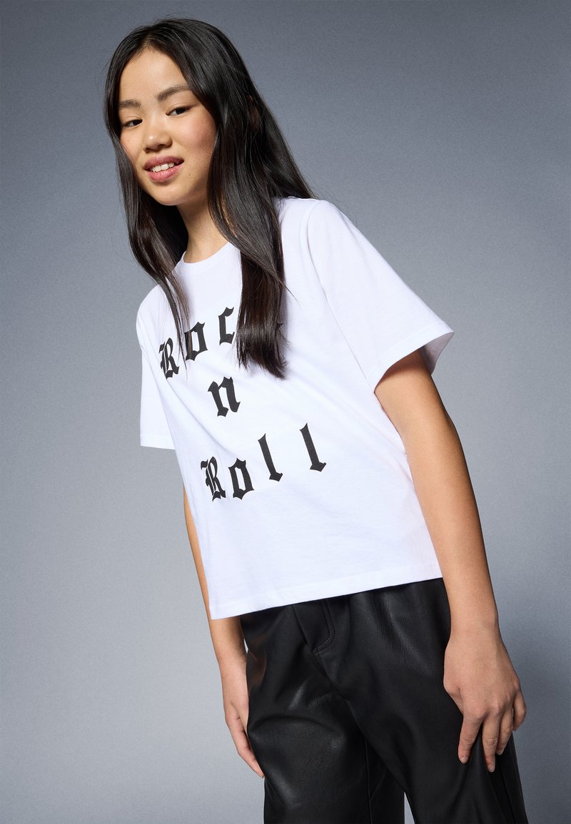 Bílý bavlněný tričko s krátkými rukávy, s výrazným černým textem "Rock n Roll" v gotickém písmu. Model je kombinován s černými koženými kalhotami.