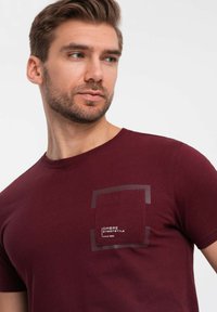 Bordeauxfarbenes T-Shirt mit kurzen Ärmeln aus Baumwolle. Verfügt über eine rechteckige aufgesetzte Tasche mit einem Logo und einem runden Ausschnitt. Glatte Textur.