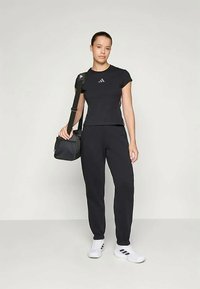 Naine mustas Adidas'i sportides ja valgetes tossudes hoiab musta õlakotti tähnilise tausta ees.