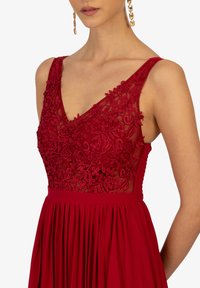 Rotes ärmelloses Kleid mit V-Ausschnitt, verziert mit aufwendiger Blumenstickerei, durchscheinendem Oberkörperdetail und plissiertem Rock. Subtile Textur und Glanz.