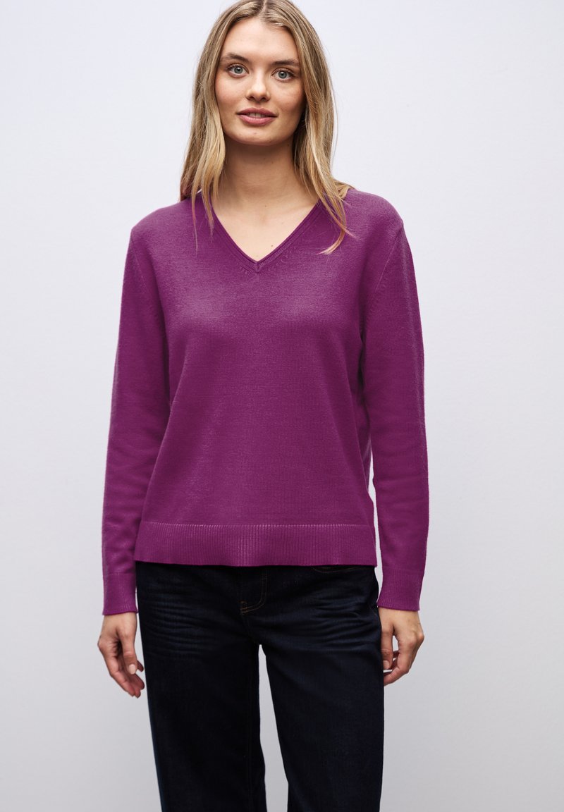 Pull V en violet en matière douce, avec des manches longues et unOurlet côtelé, associé à un jean foncé pour le contraste.