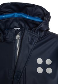 Giacca impermeabile blu navy con motivo a pois argentati, zip frontale, chiusure in velcro e fodera interna blu.