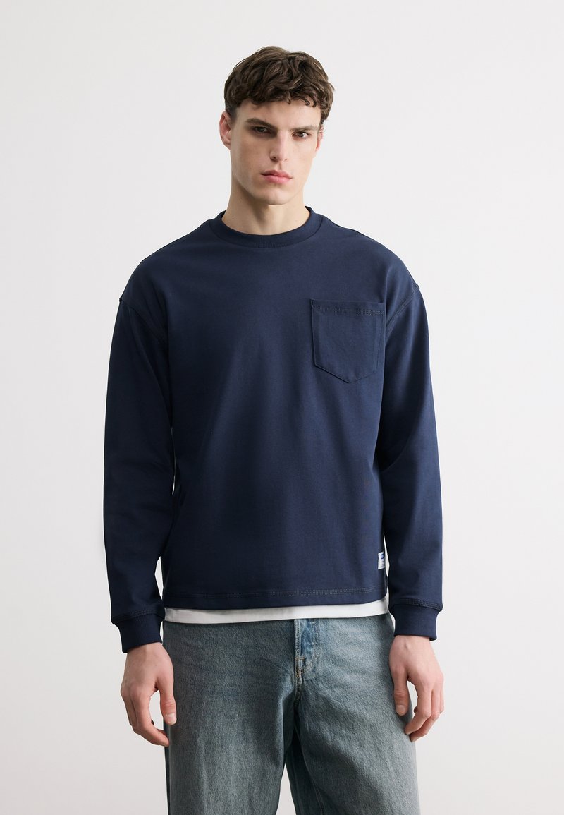 Νέος άνδρας με μακρυμάνικο μπλε πουκάμισο με τσέπη στο στήθος και ανοιχτόχρωμο τζιν, που στέκεται μπροστά σε απλό φωτεινό φόντο.