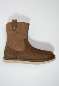 Ruskean mokkanahkainen nilkkabooti pyöreällä kärjellä, ommellulla yksityiskohdalla ja vetolenkillä. Pohja on beige, mikä kontrastoi tummempaan päälle.