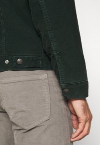 Vihreä corduroypuku, jossa on nappiriipukset, yhdistettynä vaaleanharmaisiin corduroy-housuihin, joissa on takatasku. Kankaan rakenne ja vankat yksityiskohdat.