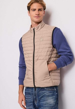 Giovane uomo con gilet trapuntato beige sopra una camicia blu a maniche lunghe e jeans blu, in piedi con una mano in tasca contro un muro semplice.