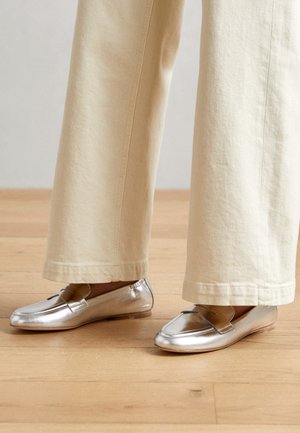 Zilverkleurige metallic platte schoenen met een gladde textuur, een afgeronde neus en een decoratieve riemdetail, gecombineerd met lichtbeige broek met brede pijpen.