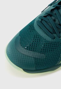 Teal sportsko med en mesh-overdel, gummidetaljer og en struktureret syntetisk tåhætte. Snørebånd og perforerede detaljer synlige.
