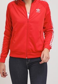 Červená sportovní bunda Adidas s bílými třemi pruhy na rukávech, zipem vpředu a dvěma bočními kapsami. Hladká tkanina s přiléhavým střihem.