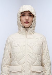 Donna che indossa un giubbotto trapuntato color crema con cappuccio regolabile, zip frontale e tasche a pattina, rivolta verso l'avanti su uno sfondo neutro.